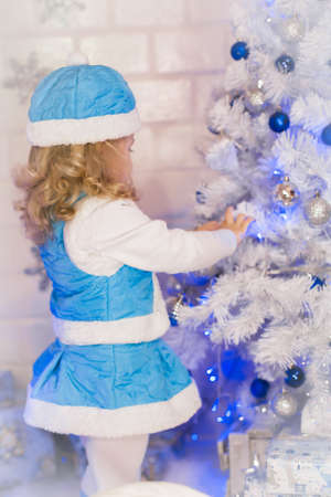 the little fair-haired curly girl in a suit of the Snow Maiden decorates a white Christmas treeの写真素材