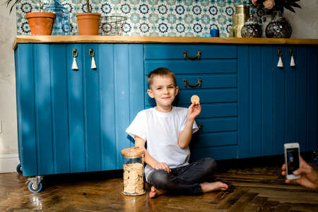 A boy in a white t-shirt is sitting on the floor. Isolated against a blue kitchen background.The boy is eating and holding a bowl of cookiesの写真素材