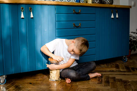A boy in a white t-shirt is sitting on the floor. Isolated against a blue kitchen background.The boy is eating and holding a bowl of cookiesの写真素材