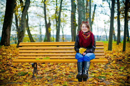 young beautiful woman sitting on a bench with yellow leaves in her hands, its autumn, she is laughingの写真素材