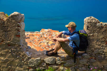 a traveller sitting on the stone fence above town Cefalu in Sicily, he is looking down at it