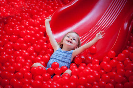 happy cute laughing little girl riding a slide in red plastic ballsの写真素材