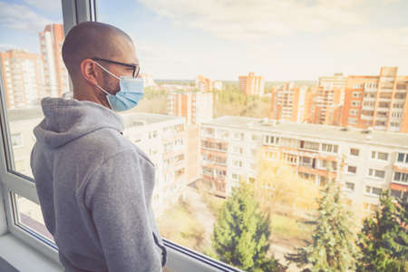 Man in protective medical mask looking through the window. Isolation at home for self quarantine. Concept home quarantine, prevention COVID-19. Coronavirus outbreak situationの写真素材