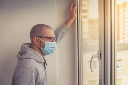 Man in protective medical mask leaning against the window. Isolation at home for self quarantine. Concept home quarantine, prevention COVID-19. Coronavirus outbreak situationの写真素材