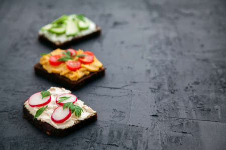 Healthy vegetarian rye bread toasts with cottage cheese, hummus, avocado, raddish and tomato. Veggie diet. Black background, close-up, copy-spaceの写真素材