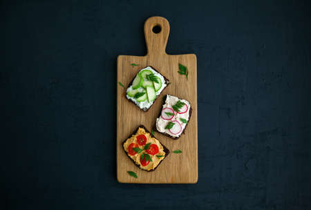 Healthy vegetarian rye bread toasts with cottage cheese, hummus, avocado, raddish and tomato. Wooden board on a black background, copy-space, flat layの写真素材