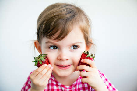 Adorable little girl holding two big strawberries near her cheeksの写真素材