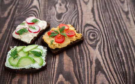 Vegetarian rye bread toasts with cottage cheese, hummus, avocado, raddish and tomato on a wooden...