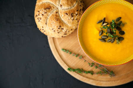Close-up tasty vegan homemade pumpkin soup, served with seeds, herbs and bun, on a black background. Top view, copy-space.の写真素材