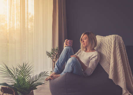 pensive woman drinking coffee and looking through window while sitting on a bean bag chair at homeの写真素材