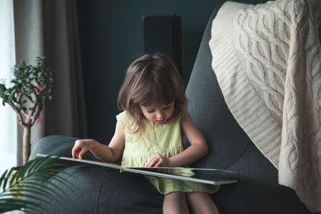 Little cute baby girl reading a book at home sitting on a bean bag chairの写真素材