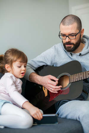 Little girl showing her father a song on a phone while he is playing guitar for herの写真素材