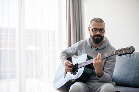 Bearded man playing guitar sitting on a sofa at home. Hobby concept.の写真素材
