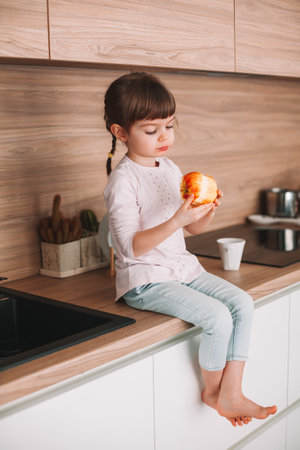 Cute little girl eating red apple sitting on a kitchen surface. Healthy eating concept.の写真素材