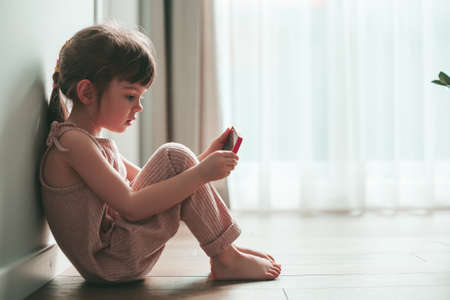 Cute little girl with mobile phone sitting alone on a floor. She is playing games or watching cartoons on smartphone. Digital generation and phone addiction concept.の写真素材