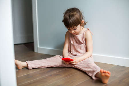 Cute little girl with mobile phone sitting alone on a floor. She is playing games or watching cartoons on smartphone. Digital generation and phone addiction concept.の写真素材