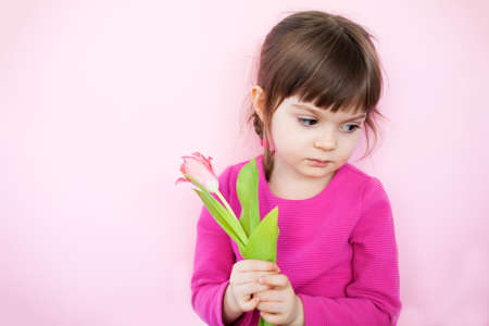 Sad little girl in pink dress holding pink tulip on a pink background