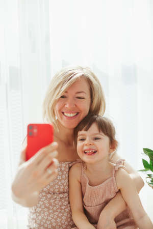 Cute little girl and her young mother are sitting together and hugging, taking selfies on a smart phone and smilingの写真素材