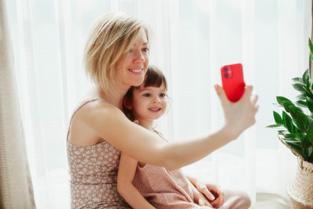 Young woman and her pretty little daughter are sitting together and hugging, taking selfies on a smart phoneの写真素材