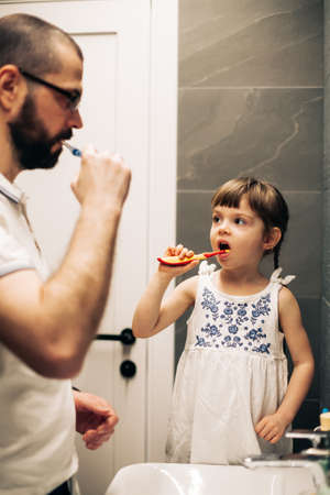 Bearded father and his little daughter brushing teeth in a bathroom and looking at each other. Man teaching his daughter how to brush teeth.の写真素材