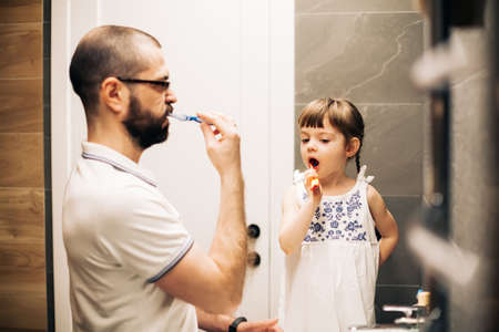 Bearded father and his little daughter brushing teeth in a bathroom. Man teaching his daughter how to brush teeth.の写真素材