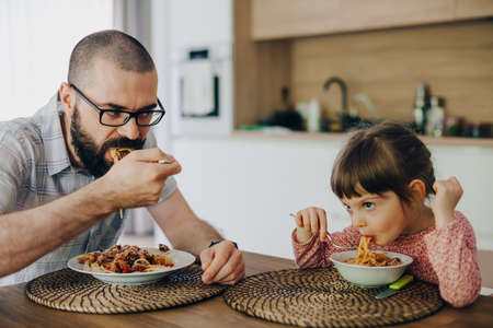 Cute little girl and her father eating spaghetti with sauce sitting in a kitchen at homeの写真素材