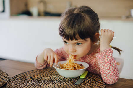 Cute little girl eating spaghetti with sauce sitting in a kitchen at homeの写真素材
