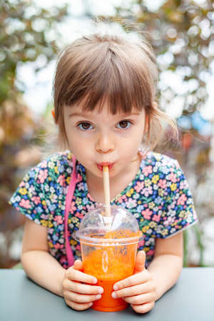 Cute 4 years old girl drinking fresh carrot juice with drinking straw seating outsideの写真素材