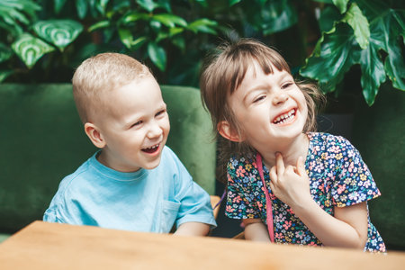 Two adorable laughing kids - friends or brother and sister - sitting indoors