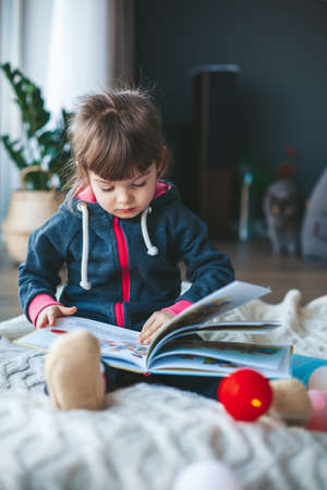 Cute little girl reading a book sitting on a woolen blanket near the window. it's winter, it's snowy and cold outside.の写真素材