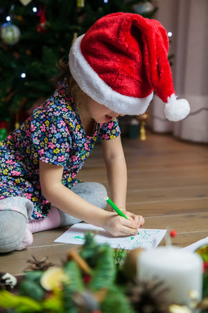 Cute little girl in a red cap writing a letter to Santa Claus near Christmas tree at home. Merry Christmas and Happy Holidays!の写真素材