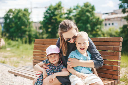 Young woman with two kids - a boy and a girl of 3-4 years, sitting on a park bench. Happy young mother with children hugging and playing fool. Happy family spending time together outside.の写真素材