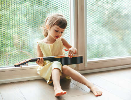 Cute little girl 4-5 years old singing and playing ukulele, sitting near the big window. Beautiful child learning to sing and to play a musical instrument.の写真素材