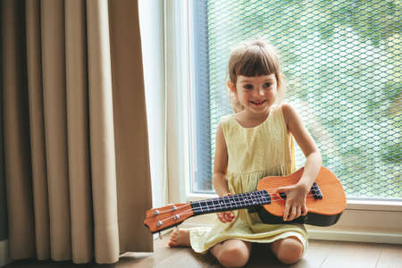 Cute little girl holding ukulele, sitting near the big window. Beautiful child learning to play a...