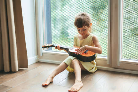 Cute little girl 4-5 years old playing ukulele, sitting near the big windowの写真素材