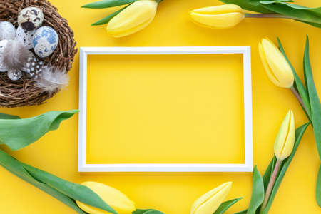 Easter holiday background with quail nest with eggs, photo frame and tulip flowers on a yellow...