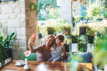 Blonde woman and her little daughter using smartphone for taking selfie in the cafeの写真素材