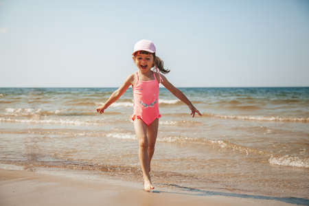 Happy adorable little girl in pink swimsuit having fun, running on the beachの写真素材