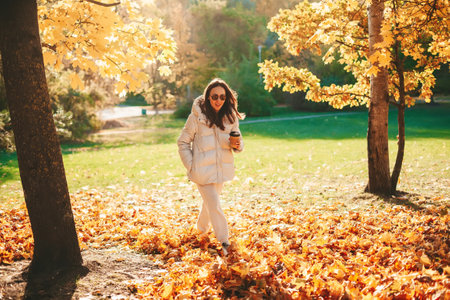 Young woman in white streetwear walking in the autumn park, drinking coffee from disposable coffee...