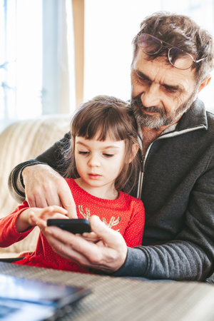 Senior man 60-65 years old and his little granddaughter using a smartphone together, sitting on a sofa. Two generations learning how to use new technologies.の写真素材