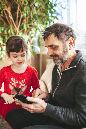 Senior man 60-65 years old using a smartphone, his little 4-5 years old granddaughter looking at him