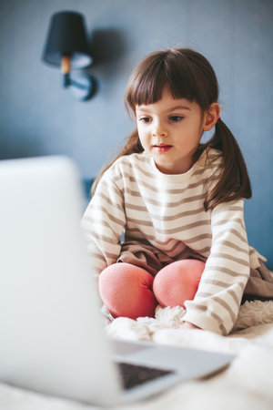 Cute little girl sitting on a bed at home and watching a video on a laptopの写真素材