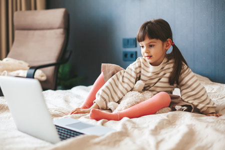 Adorable little girl sitting on a bed in a bedroom and watching something interesting on a laptop