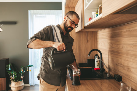 Bearded man preparing a morning black tea in the kitchen. Pouring boiling water from electric kettle into a teapot.の写真素材