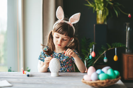 Little girl with bunny ears sitting at the table painting gypsum Easter eggの写真素材