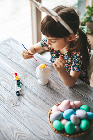 Top view little girl with bunny ears sitting at the table painting Easter egg, made of gypsumの写真素材