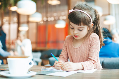 Cute little girl writing something in a notebook sitting in a cafe. Kindergarten children education, back to school, preschool child study at home concept.の写真素材