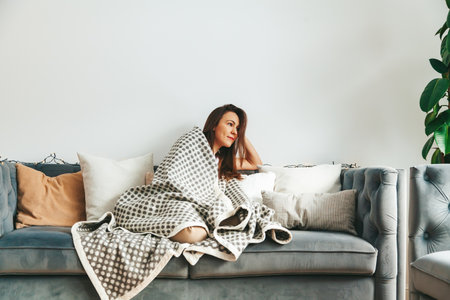 A cozy scene: a woman sitting on the couch, wrapped in a warm blanketの写真素材