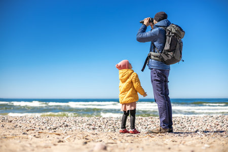 A tourist man standing on the seashore, using binoculars to observe the coastal landscape. His little daughter standing nearby.の写真素材