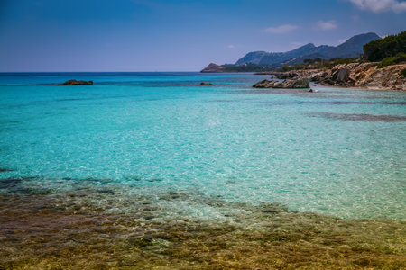 A serene coastal scene from Mallorca, the largest island of the Balearic Islands in the Mediterranean Sea. In the foreground, there is a clear turquoise sea with visible rocks beneath the shallow waterの写真素材